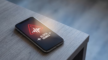 Teléfono móvil sobre mesa de madera. Pantalla encendida con "ALERTA SÍSMICA", icono de onda sísmica y triángulo de advertencia rojo.
