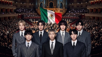 Siete hombres con traje oscuro y corbata posan en primer plano frente a una orquesta, un público en palcos y una gran bandera de México.