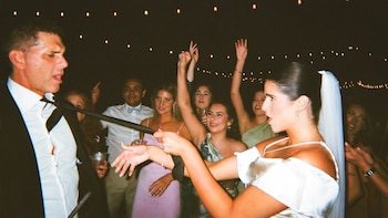 Hombre en traje y mujer con vestido de novia que tira de su corbata, rodeados de personas festejando bajo luces de guirnalda en la oscuridad