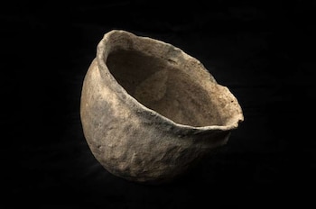 Vaso de cerámica antiguo, de color marrón claro, con forma semiesférica y borde irregular, sobre un fondo negro liso