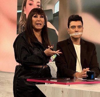 Mujer de cabello oscuro y hombre con cinta que dice "BOZAL" sobre la boca, sentados en un estudio de televisión con pantallas y escritorio