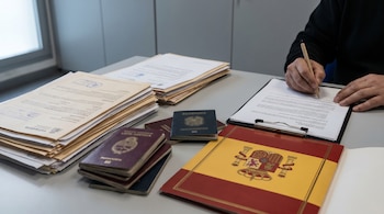 Mesa de oficina con documentos de extranjería y nacionalidad. Una mano sostiene un bolígrafo sobre un documento. Pasaportes latinoamericanos y una carpeta con el escudo de España son visibles.