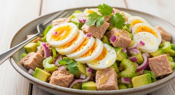 Primer plano de una ensalada fresca con trozos de atún, rodajas de huevo cocido, cubos de aguacate, apio y cebolla morada en un plato con un tenedor.