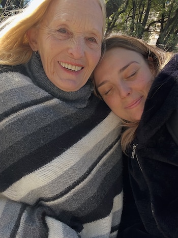 Dos mujeres vestidas con abrigo de invierno al aire libre; una sonríe hacia la cámara y la otra apoya la cabeza con los ojos cerrados