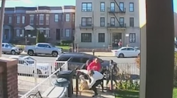 Dos personas, una con camisa roja y otra blanca, forcejean junto a un automóvil negro y una valla en una calle urbana de Brooklyn con edificios al fondo