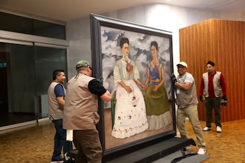 Cuatro hombres mueven un gran cuadro enmarcado que representa la obra "Las Dos Fridas", en el interior de un edificio con paneles de madera y cristal