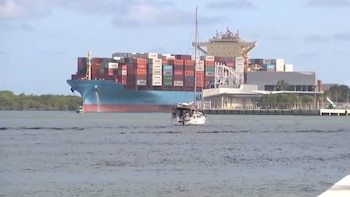 Un buque portacontenedores azul de gran tamaño, cargado con contenedores de colores, navega en el agua, con un velero blanco en primer plano y un muelle a la derecha
