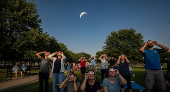 Vista de un grupo de personas en un parque, mirando hacia arriba con gafas de eclipse. El cielo está despejado y se observa un eclipse solar parcial en forma de media luna.