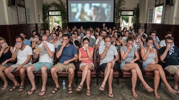 Grupo de personas sentadas en butacas de cine cubriendo sus narices con pañuelos y manos; una pantalla proyecta imágenes, palmeras a los costados.