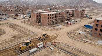 Sitio de construcción con edificios en obra, dos excavadoras, trabajadores instalando tuberías de hormigón, caminos de tierra y un paisaje de colinas con viviendas