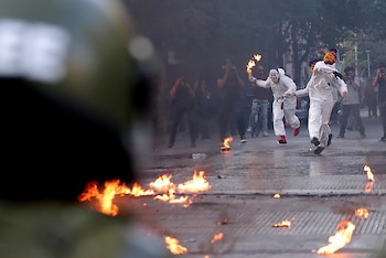 Vista desde detrás de un casco borroso de dos figuras con trajes blancos corriendo sobre una calle con llamas. Uno lleva una máscara de payaso y un cóctel Molotov