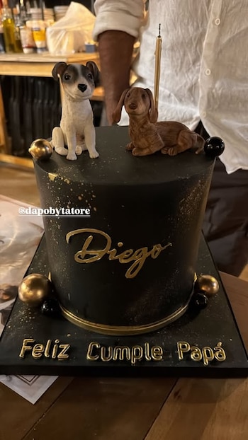 Torta de cumpleaños negra con decoración dorada, dos figuras de perros, una vela, y los textos 'Diego' y 'Feliz Cumple Papá'
