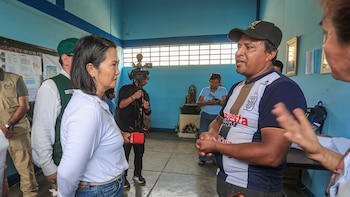 Keiko Fujimori, un hombre con gorra negra y camiseta de fútbol, un camarógrafo y más personas en una sala con paredes azules y piso de concreto
