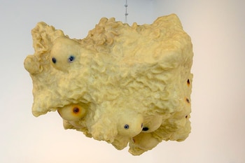 Escultura suspendida de forma irregular en tono amarillo pálido, con superficie texturizada y varios ojos protuberantes con iris oscuros