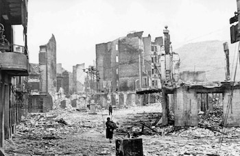 Imagen en blanco y negro de una calle destruida en Guernica; se ven escombros, edificios derruidos y dos personas caminando por el centro de la escena