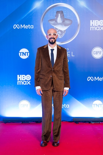 Emiro Navarro, un hombre calvo con barba y gafas blancas, de pie en una alfombra roja, vistiendo un traje marrón con patrón en espiga y corbata azul