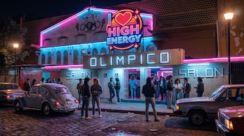 Entrada del ex Balneario Olímpico iluminada con neones rosa y azul, letrero 'I Love High Energy'. Personas se congregan en la entrada; dos autos clásicos, incluido un Vocho, están estacionados.