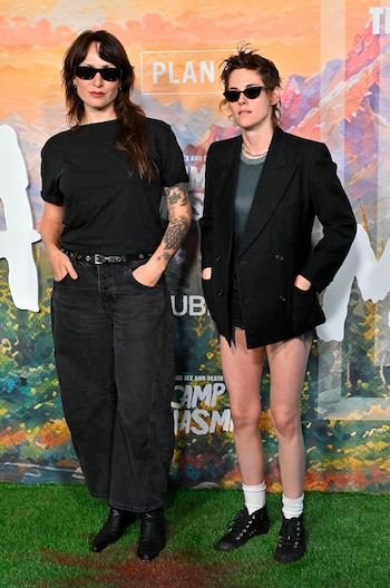 Dos mujeres con gafas de sol. Una viste camiseta y pantalones negros. La otra lleva chaqueta negra, shorts, calcetines y zapatillas. Fondo con diseño de paisaje