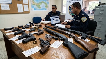 Pistolas, revólveres y escopetas con etiquetas sobre una mesa frente a dos policías trabajando en computadoras.