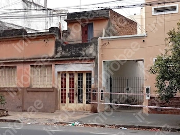 Fachada de una casa de tonos claros con ventanas, una puerta de madera y reja metálica en la entrada, delimitada por cinta de seguridad rojiblanca