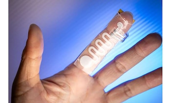 Mano abierta con la palma hacia arriba, dedo índice con un dispositivo transparente flexible adherido que muestra patrones blancos ondulados y puntos circulares, fondo azul borroso
