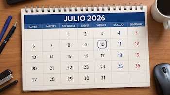 Un calendario de escritorio de Julio de 2026 con el día 10 en un círculo azul. Hay dos bolígrafos, una grapadora, un ratón de ordenador y una taza.