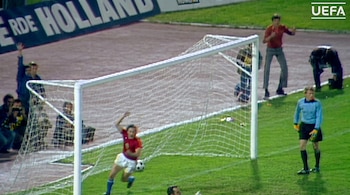 Un jugador de fútbol con camiseta roja y amarilla junto al arco, un portero con camiseta azul, una pelota en el césped, público y vallas publicitarias.
