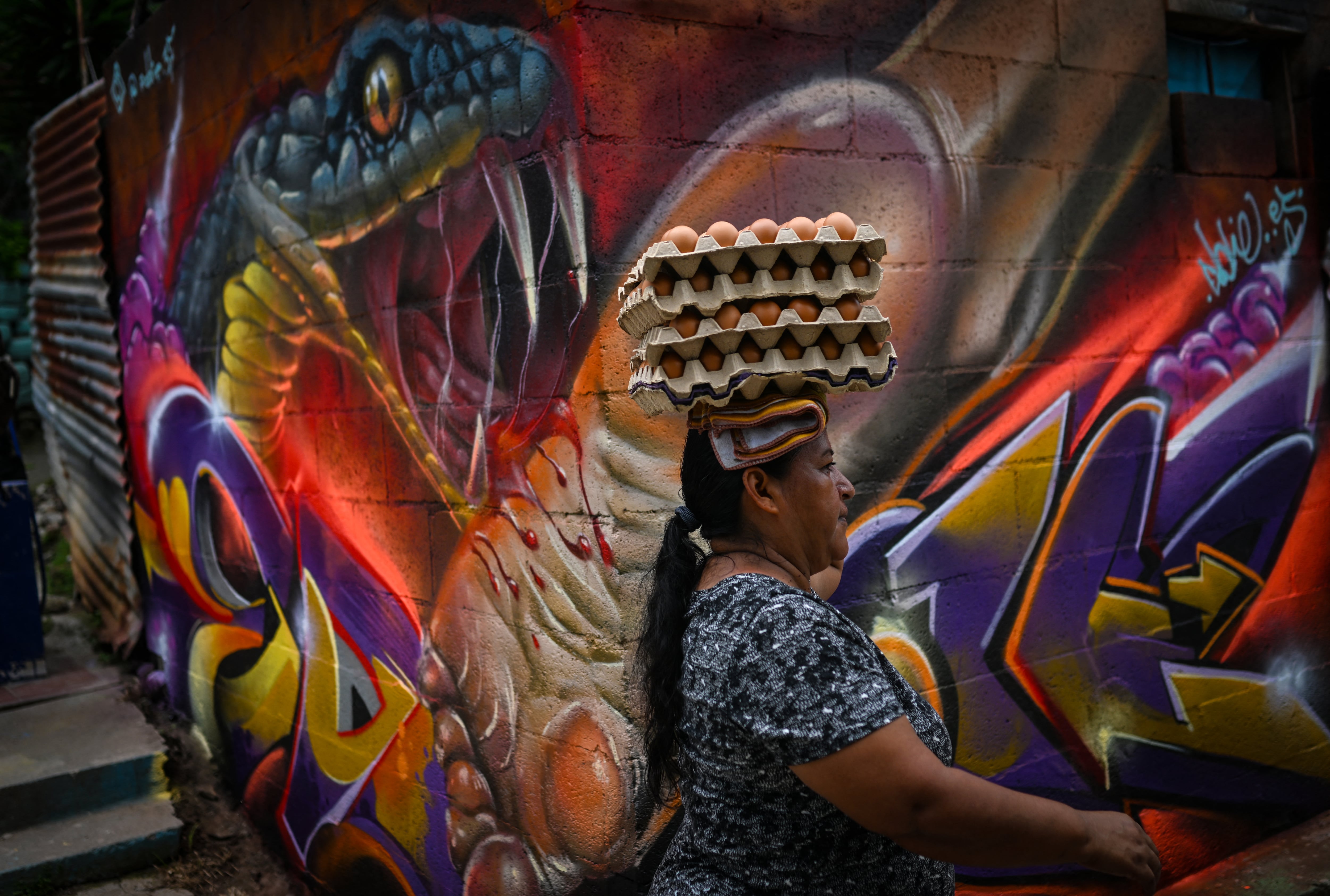 Una mujer que lleva una bandeja de huevos sobre la cabeza pasa junto a un mural creado como parte de un festival de arte callejero en la comunidad 10 de Octubre de San Marcos, El Salvador, el 2 de octubre de 2026. El proyecto artístico busca transformar la identidad de una comunidad históricamente afectada por la violencia de las pandillas. (Foto de Marvin RECINOS / AFP)