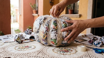 Manos que envuelven con cinta adhesiva una bolsa con papel sobre una mesa con mantel bordado, tijeras y revistas.