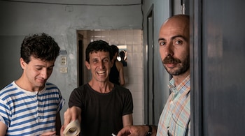 Tres hombres en el interior de una vivienda; uno lleva camiseta a rayas, otro ríe con un rollo de cinta y el tercero tiene bigote.