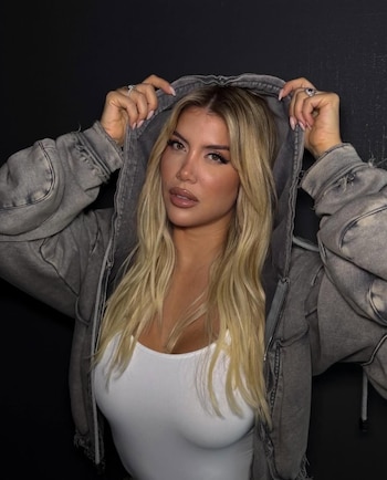 Wanda Nara con cabello rubio largo, posa frente a un fondo oscuro, llevando una chaqueta con capucha gris y una camiseta blanca. Sostiene la capucha con ambas manos