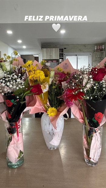 Tres floreros con ramos de flores variadas sobre una mesa de madera junto con el mensaje Feliz Primavera en la parte superior
