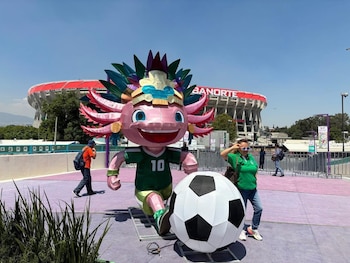 Estatua gigante de ajolote rosa con camiseta verde número 10, pateando un balón de fútbol enorme. Una mujer posa a su lado. Estadio CDMX al fondo
