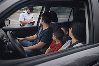 Familia con dos adultos y dos niños dentro de un automóvil gris, vista desde la ventanilla del acompañante. Un policía de tránsito escribe una multa afuera.