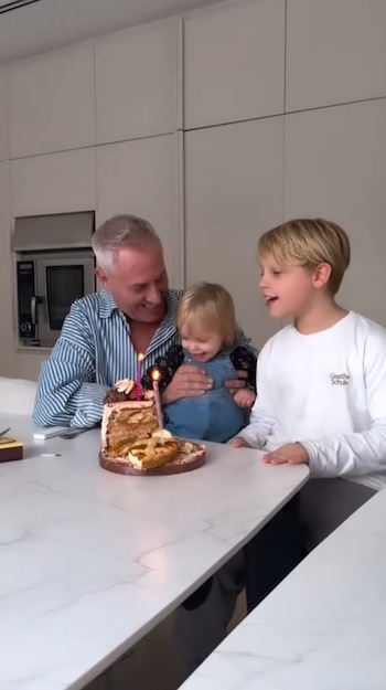 Marley sonríe sosteniendo a un bebé que ríe, junto a un niño mayor. Una torta con dos velas encendidas y un trozo de tarta están sobre la mesa blanca