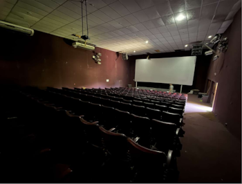 Interior de un teatro o centro de artes escénicas con butacas vacías, una gran pantalla al fondo y luminarias en el techo y paredes, poco iluminado