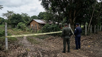 Dos agentes de policía de espaldas vigilan la entrada a una finca rural, acordonada con cinta amarilla de 'escena del crimen', con una casa rústica y árboles frondosos al fondo.