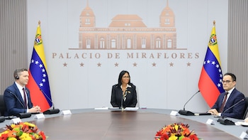 Delcy Rodríguez, con traje oscuro y gafas, preside una mesa de reunión con dos hombres sentados a sus lados, flanqueados por banderas venezolanas en el Palacio de Miraflores