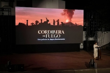 Gran pantalla muestra el título de la película 'Cordillera de Fuego' y siluetas de personas con un volcán en erupción. Mujer habla en podio con micrófono en un escenario oscuro
