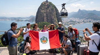 Ocho personas sostienen una bandera de Perú frente al Pan de Azúcar en Río de Janeiro, con un teleférico, maletas y la bahía al fondo.