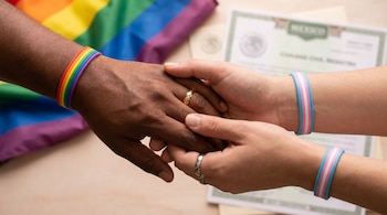 Cuatro manos entrelazadas con anillos. Una muñeca lleva pulsera arcoíris, otra pulsera trans. Fondo difuminado con bandera arcoíris y certificado de México.