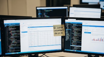 Múltiples monitores de ordenador en un entorno de oficina, mostrando código, hojas de cálculo y navegadores web, con un post-it amarillo en una pantalla que dice: 'No confiar ciegamente en la IA'.