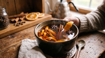 Taza de cerámica con líquido humeante, tiras de cáscara de naranja, varas de canela y anís estrellado sobre una mesa rústica.