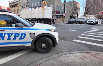 Un coche patrulla de la Policía de Nueva York aparcado en una intersección urbana con paso de peatones, comercios y vehículos al fondo
