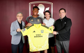 Cuatro hombres sonríen mientras sostienen una camiseta de fútbol amarilla con el logo de Adidas, el escudo del Aston Villa y la frase 'Visit Rwanda'