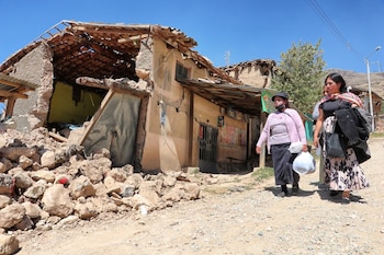 Dos mujeres caminan en una carretera de tierra frente a una casa con techo y paredes parcialmente colapsados. Hay escombros en el suelo bajo un cielo azul