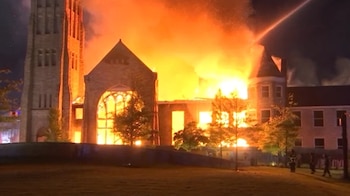 Vista nocturna del histórico Templo Clayborn, un edificio de piedra con una torre, completamente envuelto en llamas. Bomberos combaten el incendio con chorros de agua