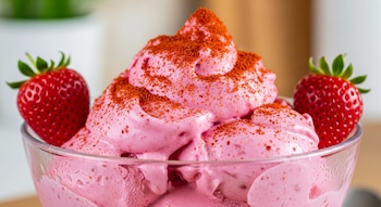 Un vaso de cristal lleno de helado de fresa rosado, cubierto con polvo de chile rojo, flanqueado por dos fresas frescas y enteras a cada lado.