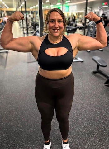 Mujer con top negro y mallas oscuras flexiona los bíceps mientras sonríe en el interior de un gimnasio