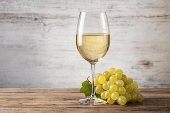 Una copa de vino blanco llena se encuentra junto a un racimo de uvas verdes con una hoja, sobre una superficie de madera y un fondo de madera clara.